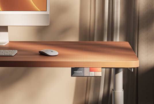Shappa electric standing desk close-up with wood-tone tabletop, mounted controller with memory presets, and white frame leg in a warm sunlit home office