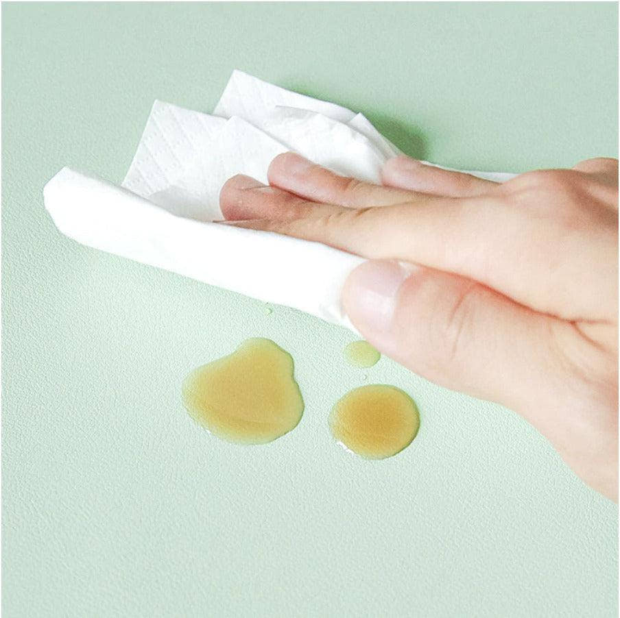Wiping a spill off the desk mat's waterproof surface with a tissue, demonstrating easy cleaning